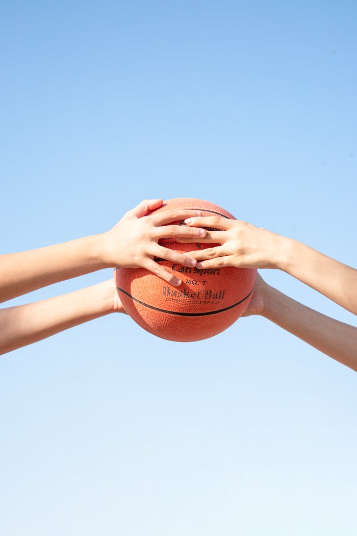 Hands of two people holding a basketball against a bright blue sky, symbolizing teamwork.